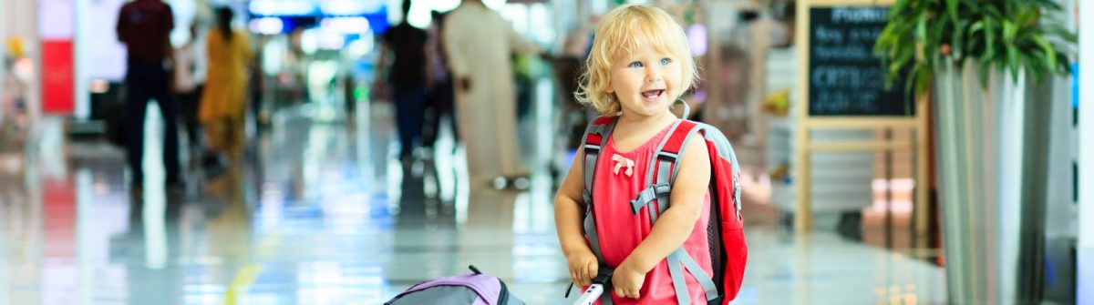 Kleinkind mit rotem Rucksack lächelt im Flughafenterminal, neben sich ein Rollkoffer. Fliegen mit Kindern leicht gemacht.