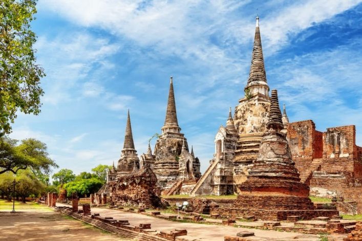 Drei spitze Pagoden der historischen Königsstadt Ayutthaya in Thailand ragen unter blauem Himmel hervor, umgeben von alten Ruinen und Grünflächen.