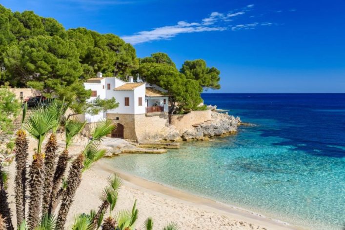 Klares türkisfarbenes Meer trifft Sandstrand, umgeben von grünen Bäumen mit weißem Haus, Top 20 Mallorca-Schönheit.