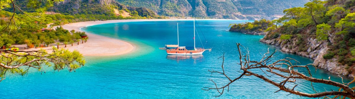 Türkiser Meeresarm umgeben von grüner Vegetation und Strand, über dem ein Segelschiff kreuzt, eingerahmt von Nadelbaumästen.