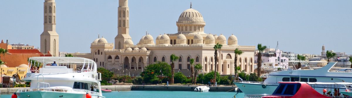 Im Vordergrund weiße und rote Boote auf blauem Wasser, dahinter eine große Moschee mit Kuppeln und Minaretten, unter klarem Himmel.