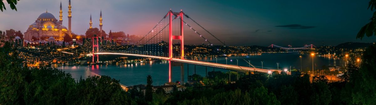 Panorama Istanbuls bei Nacht: Links die beleuchtete Süleymaniye-Moschee, zentral die glänzende Bosphorus-Brücke, rechts im Dunkeln die Bosporus-Küste.