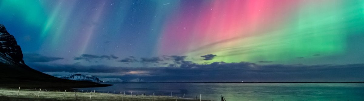 Schillernde Polarlichter in Pink und Grün zieren den Nachthimmel über einem ruhigen Meer, begleitet von der Silhouette eines Gebirges.
