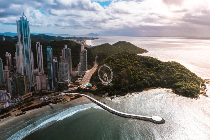 Luftaufnahme von Balneário Camboriú in Brasilien: Wolkenkratzer neben sandigem Strand, dahinter Meer, rechts bewaldete Hügel mit Riesenrad.