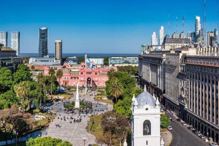 Blick auf die Plaza de Mayo in Buenos Aires an einem sonnigen Tag. Im Vordergrund Palmen und Passanten, dahinter das rosa gefärbte Casa Rosada und diverse Hochhäuser.