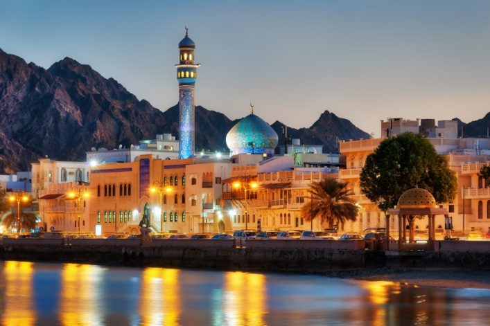 Eine Moschee mit leuchtender Kuppel und Minarett steht vor Bergkulisse in Muscat, Oman. Traditionelle Architektur umgibt sie; glänzendes Wasser im Vordergrund.