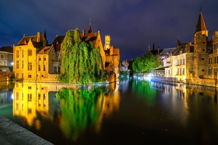 Historische Gebäude spiegeln sich in einem ruhigen Kanal bei Nacht; bunte Lichter illuminieren die Szene.