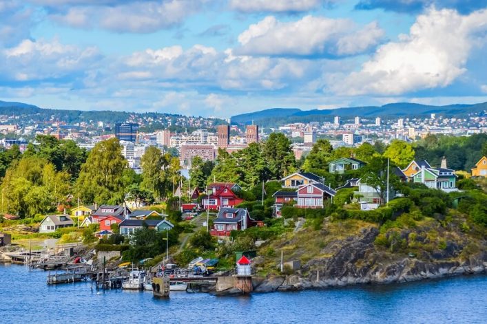 Blick auf den Oslofjord. Im unteren Teil des Bildes fließt der Fluss, darüber kommt die Küste mit Natur und Häusern. Im Hintergrund sind Oslo und Berge zu sehen. Der Himmel ist blau, aber bewölkt.