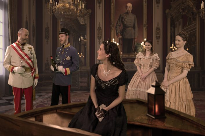 Fünf Menschen in historischen Kostümen in einem prunkvollen Raum; links zwei Männer mit Wein, rechts zwei Frauen in Kleidern, eine Frau in Schwarz sitzt in der Mitte; im Hintergrund ein großes Gemälde.
