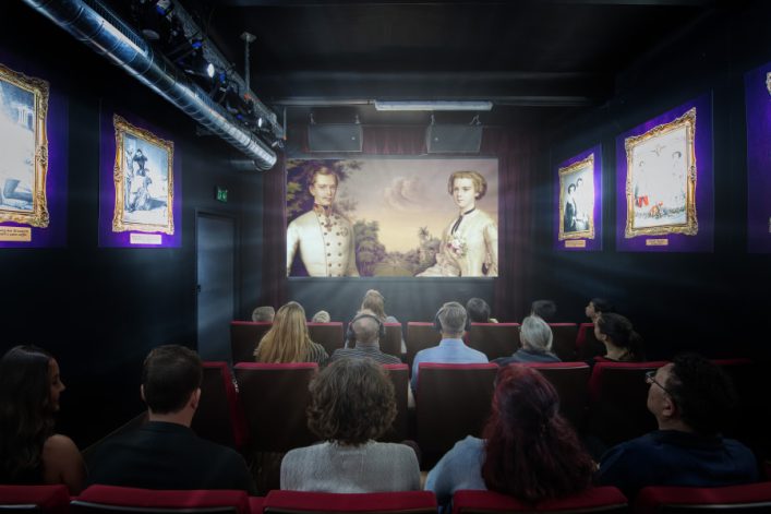 Menschen sitzen in einem kleinen Kino. Auf der Leinwand ein historisches Gemälde von zwei Figuren in edler Kleidung. An den Wänden hängen vergoldete gerahmte Bilder mit lila Hintergrund.