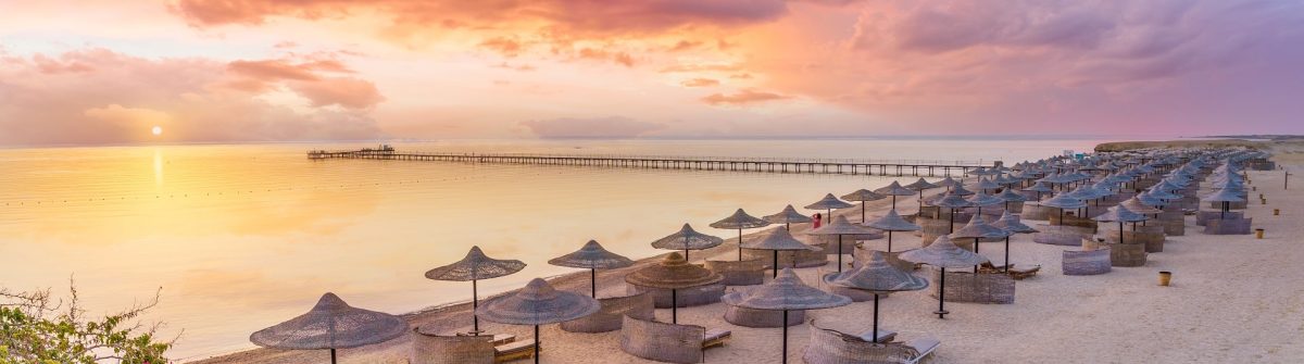 Sonnenuntergang in Hurghada: Im Vordergrund ein Strand mit geflochtenen Sonnenliegen und Strohschirmen. Ein Steg erstreckt sich ins Meer. Der Himmel ist in violetten, orangefarbenen und rosa Tönen erleuchtet.