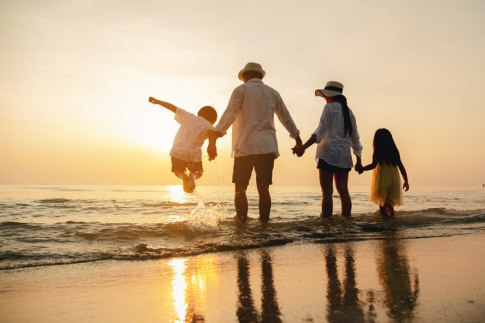 Eine Familie steht am Strand bei Sonnenuntergang. Ein Kind springt ins Meer, während die Eltern und ein weiteres Kind Hand in Hand am Wasser entlanglaufen. Packliste für eine Kreuzfahrt: Sonnenhut, lockere Kleidung.