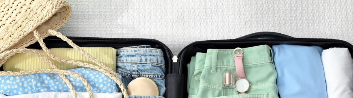 Eine geöffnete Reisetasche liegt auf einem weißen Bettlaken. In der linken Hälfte sind eine geflochtene Strohtasche, gefaltete Kleidung in Blau-, Grün- und Gelbtönen, Jeansshorts und ein Sonnenhut sichtbar. Auf der rechten Seite sind grüne Shorts mit einer rosa Armbanduhr gelegt, blaue und weiße Oberteile. Perfekte Packliste für eine Kreuzfahrt!