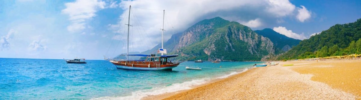 Panoramablick auf einen Strand in Antalya; ein Boot ankert im klaren, türkisfarbenen Wasser nahe dem Ufer. Rechts erstreckt sich der helle Kieselstrand, im Hintergrund grüner Wald und hohe Berge.