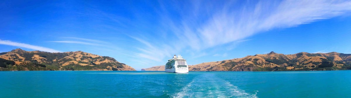 Foto eines weißen Kreuzfahrtschiffes von hinten. Im Wasser hinter dem Schiff ist weiße Gischt. Am Horizont sind rechts und links spärlich bewachsene braune Hügel zu erkennen. Der Himmel ist blau.