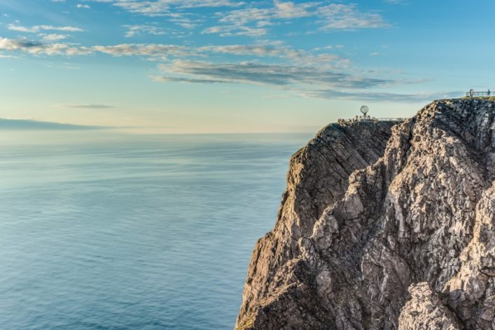 Auf der rechten Seite des Bildes liegt die Klippe des Nordkaps, oben drauf steht die Globus Statue. Links ist das Meer, oben der blaue Himmel mit wenigen Wolken.