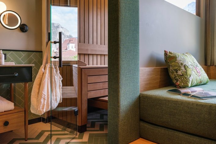 Moderne Hotelsuite mit Daybed und Glas-Sauna. Links ein Waschbecken und runder Spiegel, rechts ein grünes Sofa mit Blumenkissen, auf dem eine Zeitschrift liegt. Durch die Sauna blickt man auf Berge.