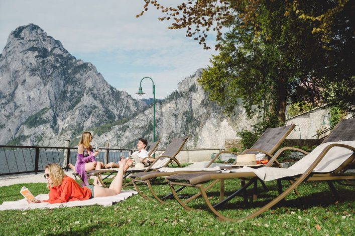 Im Bild liegt eine Person auf einer Decke im Vordergrund, liest ein Buch. Drei Liegestühle stehen rechts, teils belegt. Hinten zwei Personen mit Getränken, links Berge und Wasser, oben ein Baum mit Herbstblättern.