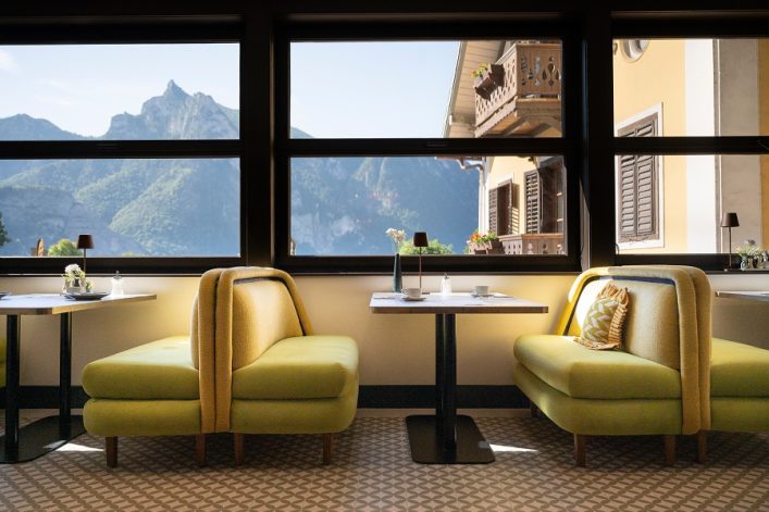 Gemütliches Café mit gelben Sofas, Tischen mit Blumen und Blick durch große Fenster auf Berge und ein gelbes Haus mit Holzbalkon. Fliesenboden und Lampen ergänzen das Ambiente.