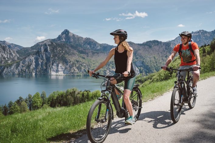 Zwei Radfahrer in Sportkleidung und Helmen fahren auf einem sonnigen Pfad im Grünen. Im Hintergrund erstrecken sich ein ruhiger See und beeindruckende Berglandschaften. Der vordere Radfahrer lächelt nach links.