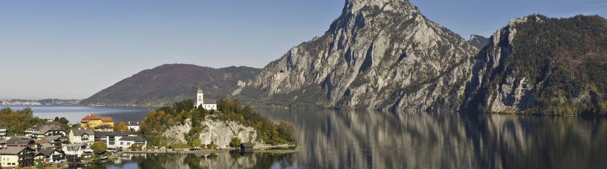 Von einem klaren See erhebt sich eine kleine Halbinsel mit einer Kirche, im Hintergrund majestätische Berge, deren Spiegelung sich im Wasser zeigt. Im Vordergrund segeln Boote entlang einer malerischen Stadt.