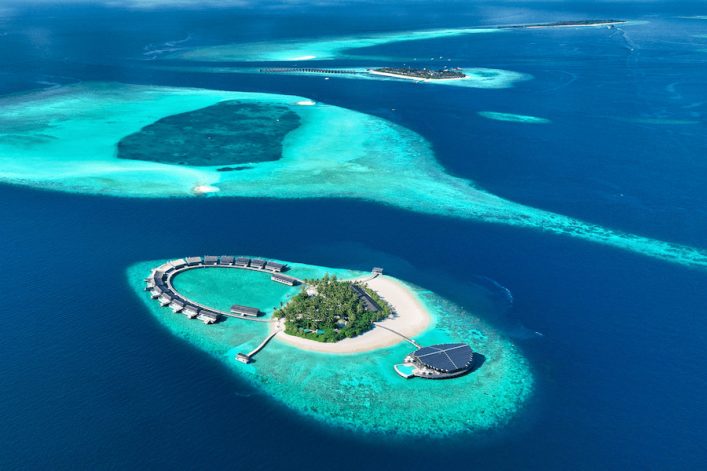 Luftaufnahme einer maledivischen Inselgruppe: Im Vordergrund eine hufeisenförmige Insel mit Wasservillen, grüner Vegetation und einer Sandbank. Umgeben vom türkisen Ozean und flachen Riffen; im Hintergrund weitere Inseln.