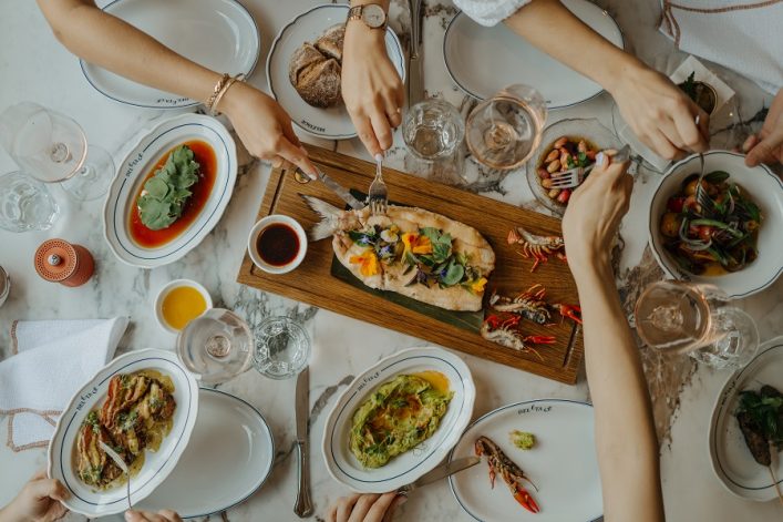 Auf einem festlich gedeckten Tisch mit Marmortischplatte greifen mehrere Hände nach verschiedenen Gerichten, darunter ein dekorierter Fisch auf einem Holztablett, Salate und Schalen mit Garnelen und Soßen.
