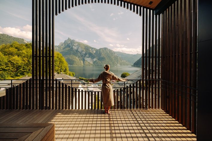 Eine Frau in einem Bademantel steht auf einer überdachten Terrasse mit Holzlamellen und blickt zum ruhigen See und zu den riesigen Bergen in der Ferne. Der Boden wirft Schattenspiele der Lamellen.