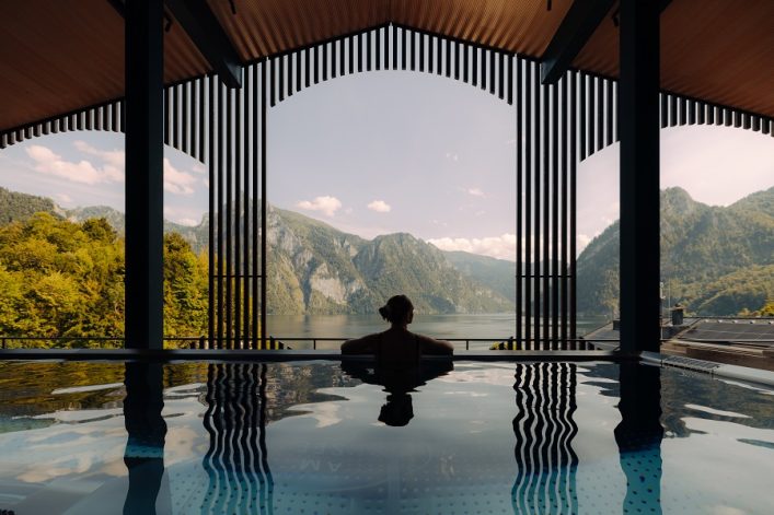 Eine Person lehnt sich am Rand eines Infinity-Pools, der durch dunkle Säulen und Holzverkleidung eingerahmt ist. Im Hintergrund erstrecken sich ein malerischer See und grüne Berge unter einem bewölkten Himmel.