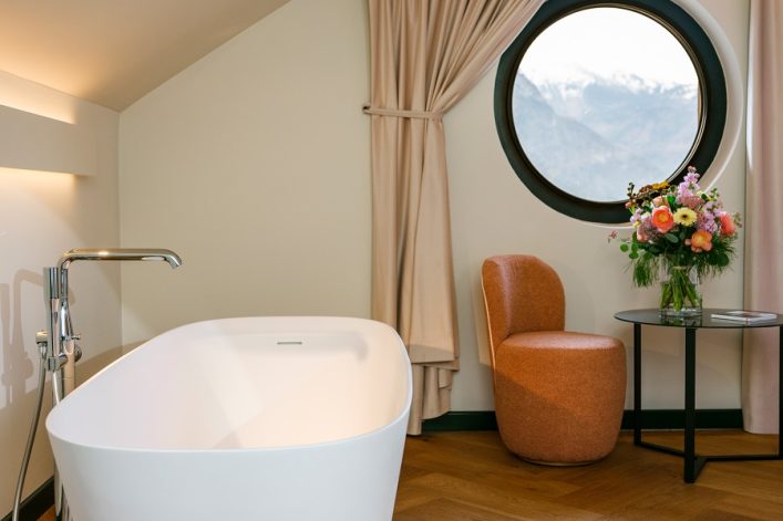 Freistehende Badewanne links, daneben ein rundes Fenster mit Bergblick, beige Vorhänge, rechts ein oranger Sessel und ein Tisch mit Blumen in einer Vase. Holzboden vervollständigt das luxuriöse Ambiente.