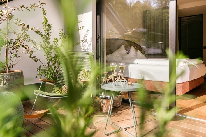 Eine gemütliche Terrasse mit Schaukelstuhl, Pflanzen und einem runden Tisch, auf dem zwei Sektgläser und eine Flasche stehen. Im Hintergrund ein großes Glasfenster mit Blick auf ein modernes Schlafzimmer.
