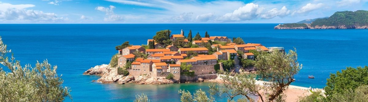 Sveti Stefan in Montenegro: Insel mit antiken Häusern und orangefarbenen Dächern, umgeben von türkisblauem Meer, unten Bäume im Vordergrund, an einem wolkigen, aber sonnigen Tag. Im Hintergrund ist die Küste sichtbar.