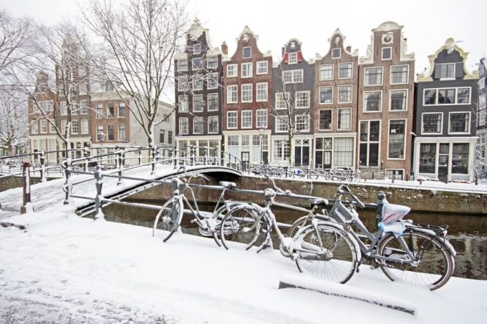 Verschneite Fahrräder lehnen an einem Geländer vor einem schneebedeckten Kanal. Dahinter stehen typische Amsterdamer Häuser mit Giebeln. Eine Brücke überspannt den Kanal, Bäume stehen kahl im Hintergrund.