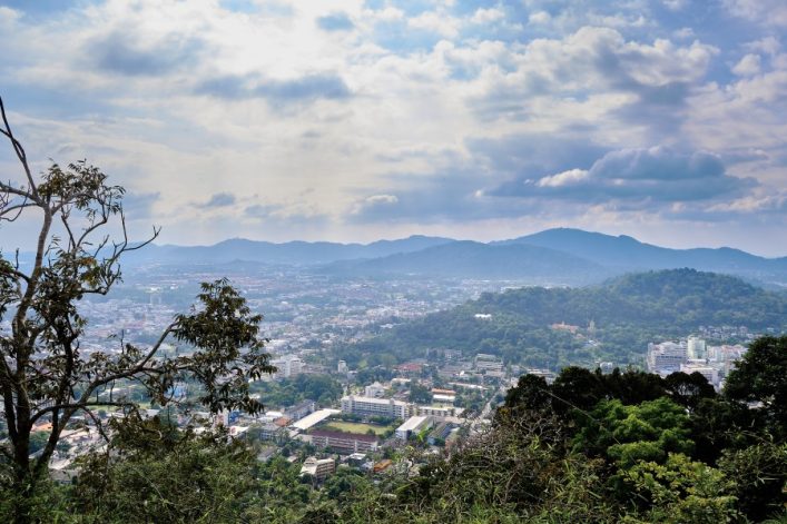 Aussichtspunkt in Phuket mit Blick auf die Stadt und bewaldete Hügel im Hintergrund. Links steht ein Baum im Vordergrund, während wolkiger Himmel die Szene überspannt.