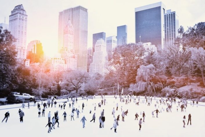 Menschen, die schlittschuhlaufen im Central Park, umgeben von kahlen Bäumen, mit der Skyline von New York im Hintergrund und Sonnenlicht, das durch die Gebäude schimmert.