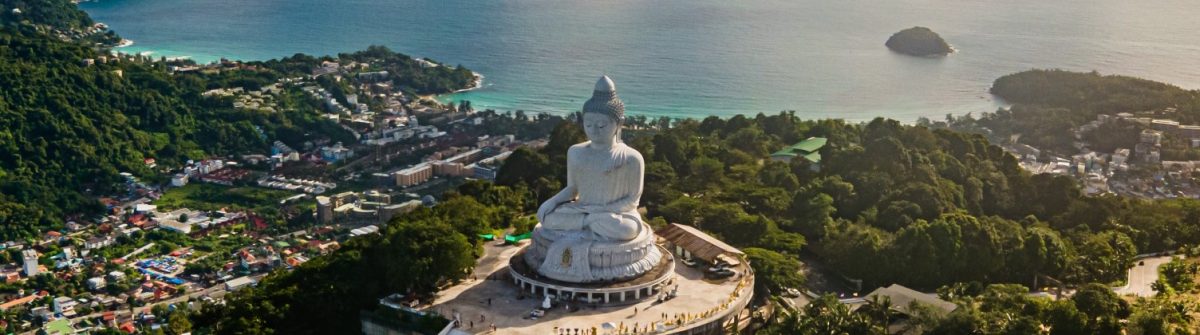 Große, weiße Buddha-Statue auf Hügel mit Blick auf bewaldete Landschaft und Küstenlinie. Der Ozean erstreckt sich im Hintergrund. Kleine Gebäude und Straßen säumen die Hänge im Vordergrund.