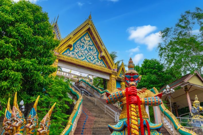 Majestätischer thailändischer Tempel mit verziertem Dach, bunte, große Wächterstatuen rechts. Treppe führt hinauf, umgeben von üppigem grünen Laub. Blauer Himmel im Hintergrund.
