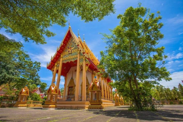 Ein reich verzierter, gold-roter Tempel steht zentral zwischen Bäumen unter klarem blauem Himmel. Im Vordergrund breitet sich eine gepflasterte Fläche aus, malerisch von Sonnenlicht erhellt.