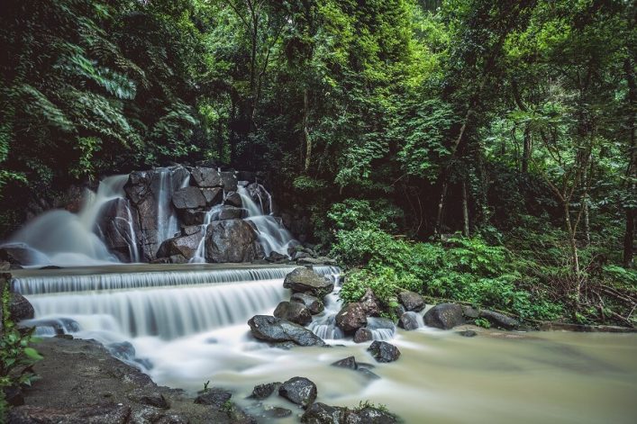 Kaskadierender Wasserfall in einem dichten, grünen Wald. Wasser strömt über große, gestapelte Felsen, umgeben von üppigem Laub und Moos. Die ruhige, milchige Flussoberfläche im Vordergrund.