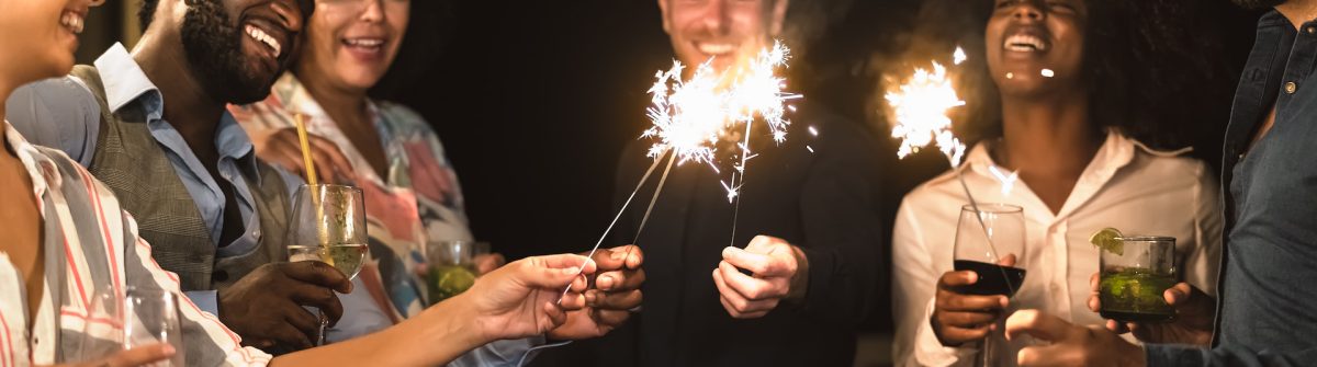 Fröhliche Menschen halten Wunderkerzen und Getränke in den Händen, feiern ausgelassen in einer Ferienwohnung über Silvester. Sie lachen und genießen die festliche Stimmung.