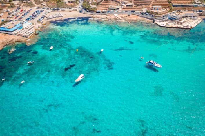 Armier Bay Beach auf Malta, türkisfarbenes Wasser mit mehreren Booten verteilt. Am oberen Rand des Bildes befinden sich Gebäude, Parkplätze und ein kleiner Hafen.