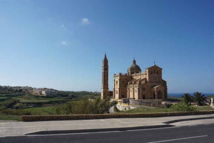 Große Basilika auf einem Hügel mit weitreichendem Blick über grasbewachsene Felder und verstreute Häuser. Im Vordergrund eine Straße, im Hintergrund blauer Himmel und das Meer.