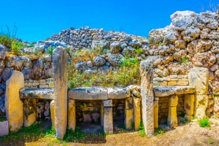 Alte Megalith-Tempelanlage mit groben, aufrechten Steinen und Büschen bewachsen. Die Anlage befindet sich vor einer Mauer aus unregelmäßig gestapelten Steinen unter klarem, blauem Himmel.