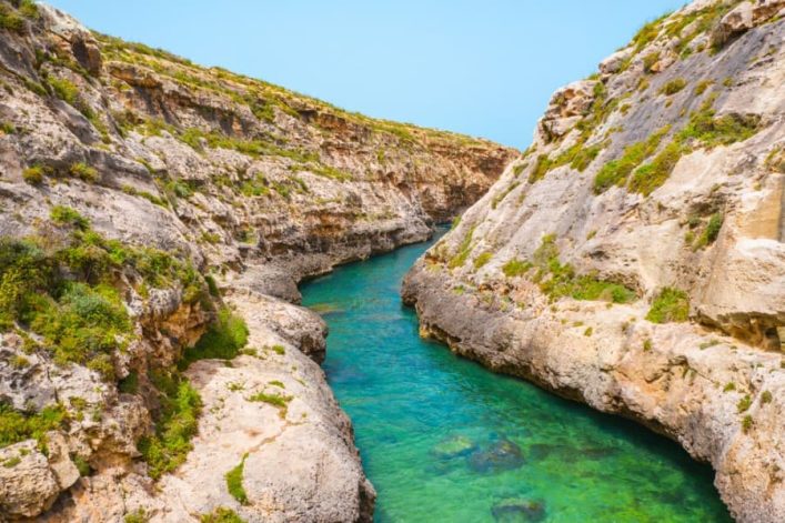 Ein enger, von hohen Felswänden umgebener Wasserkanal mit türkisfarbenem Wasser verläuft diagonal durch die Bildmitte. Über den Felsen wachsen vereinzelt Gräser. Der Himmel ist hellblau.