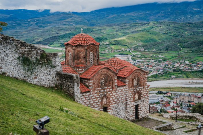 Historische Kirche mit roten Ziegeln steht auf grünem Hügel. Links alte Steinmauer, rechts Panoramablick auf Stadt und Fluss im Tal, im Hintergrund Gebirgskette unter bewölktem Himmel.